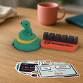 Desk Buddy Bundle: classic Pip the snake, a coral StickerCon cable dock and the ten Dev Life stickers fanned out on a wooden desk.