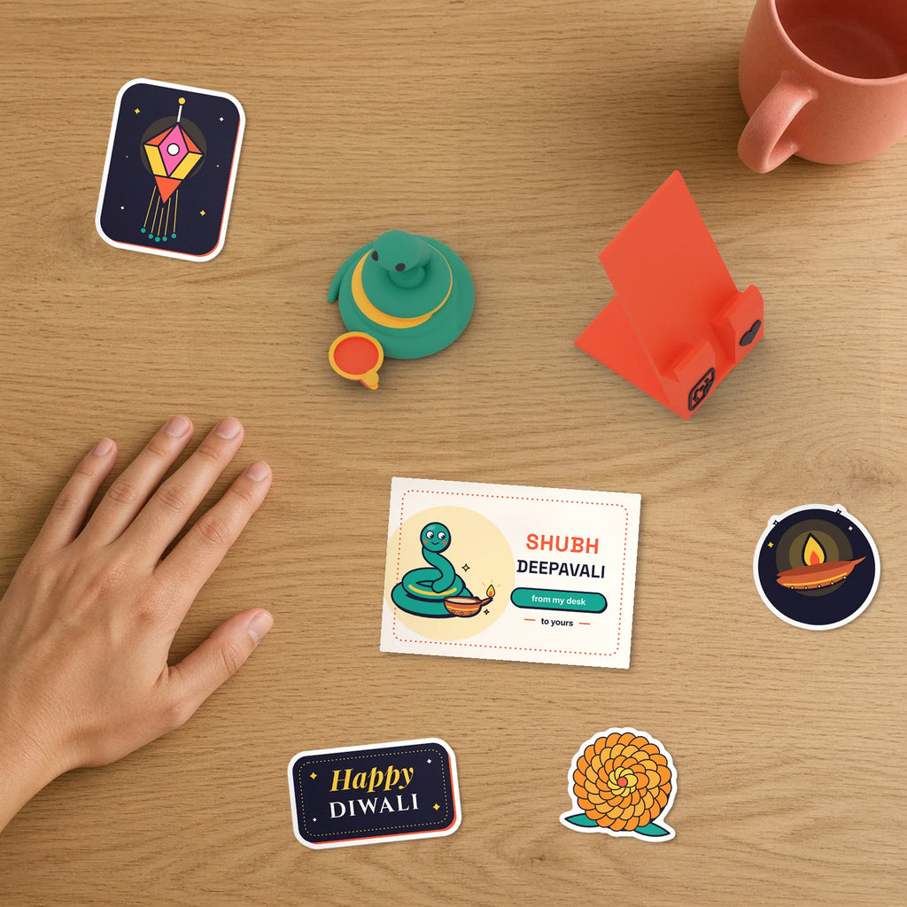 Diwali Desk Set on a desk beside a hand: Diwali Pip with a diya, phone stand, Shubh Deepavali desk card and festive stickers