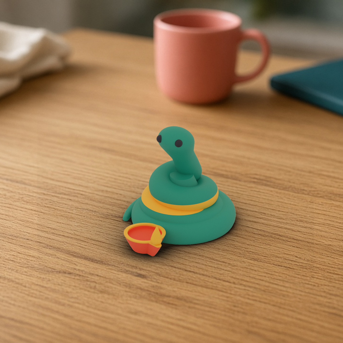 Diwali Pip: 3D printed teal snake with a yellow stripe beside a small coral clay diya, on a wooden desk.