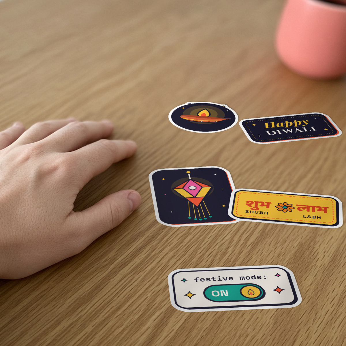 Festive Sticker Pack: five festival stickers, including a diya, a lantern and Shubh Labh, on a desk beside a hand.