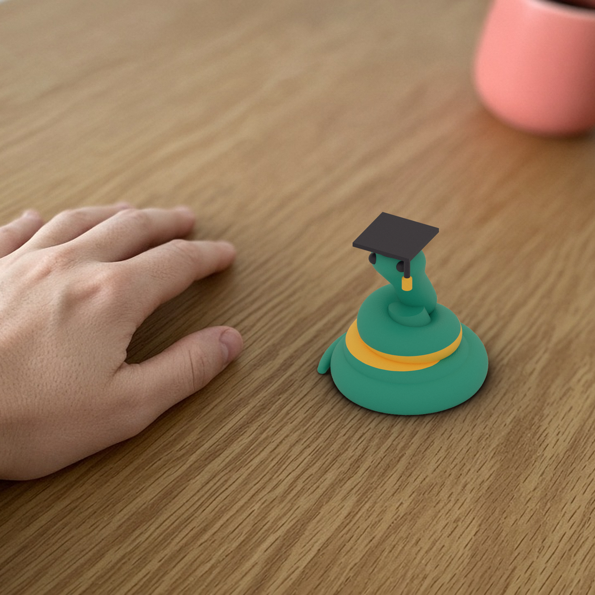 Graduate Pip: 3D printed teal snake in a graduation cap, beside a hand for scale.