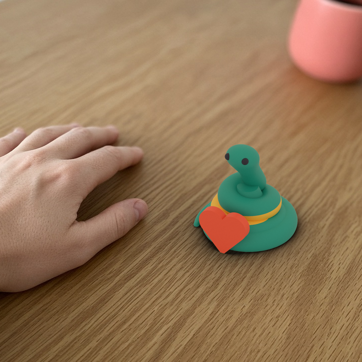 Heart Pip: 3D printed teal snake holding a coral heart, beside a hand for scale.