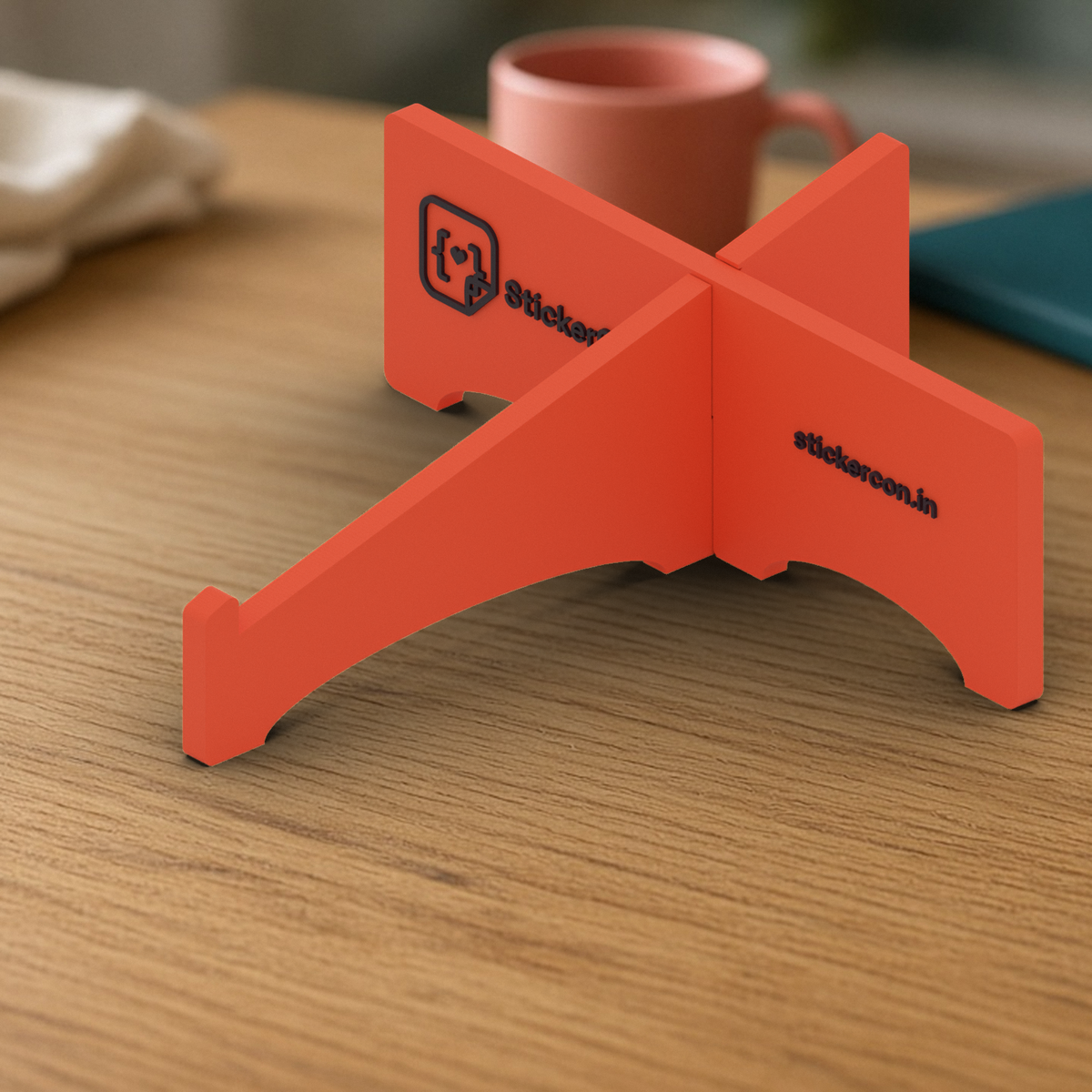 Laptop Stand: coral 3D printed cross-shaped laptop stand with the StickerCon logo, on a wooden desk.
