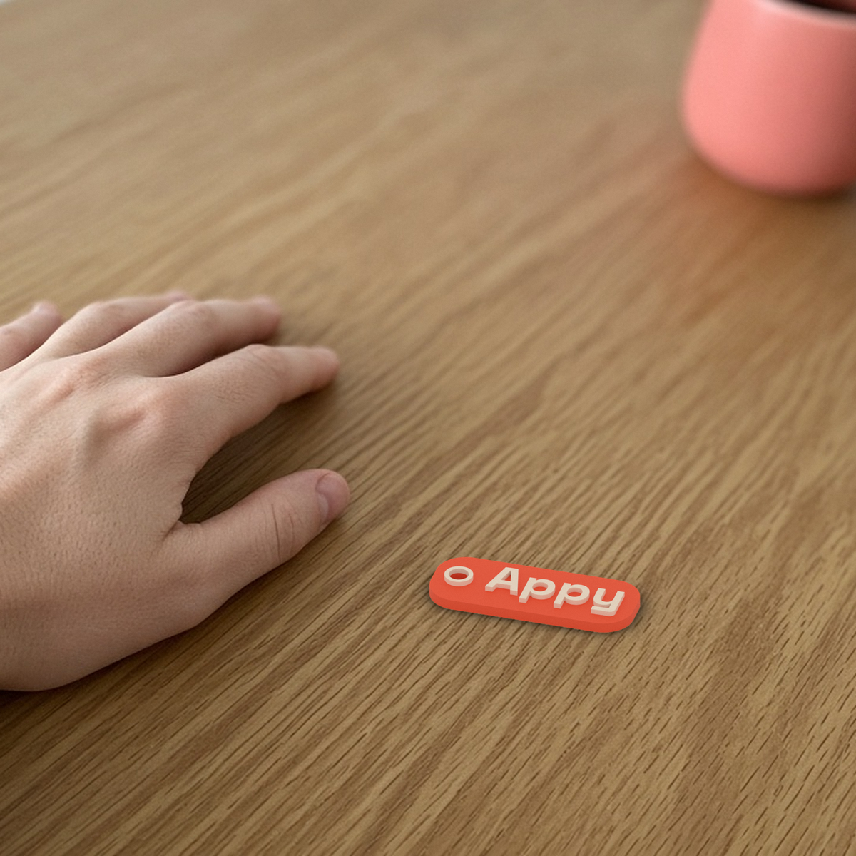 Personalised Name Keychain: coral 3D printed tag with the sample name Appy lying flat on a desk beside a hand.