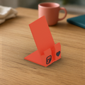 StickerCon Phone Stand: coral 3D printed phone stand with a cable slot, the StickerCon logo and a heart, on a wooden desk.