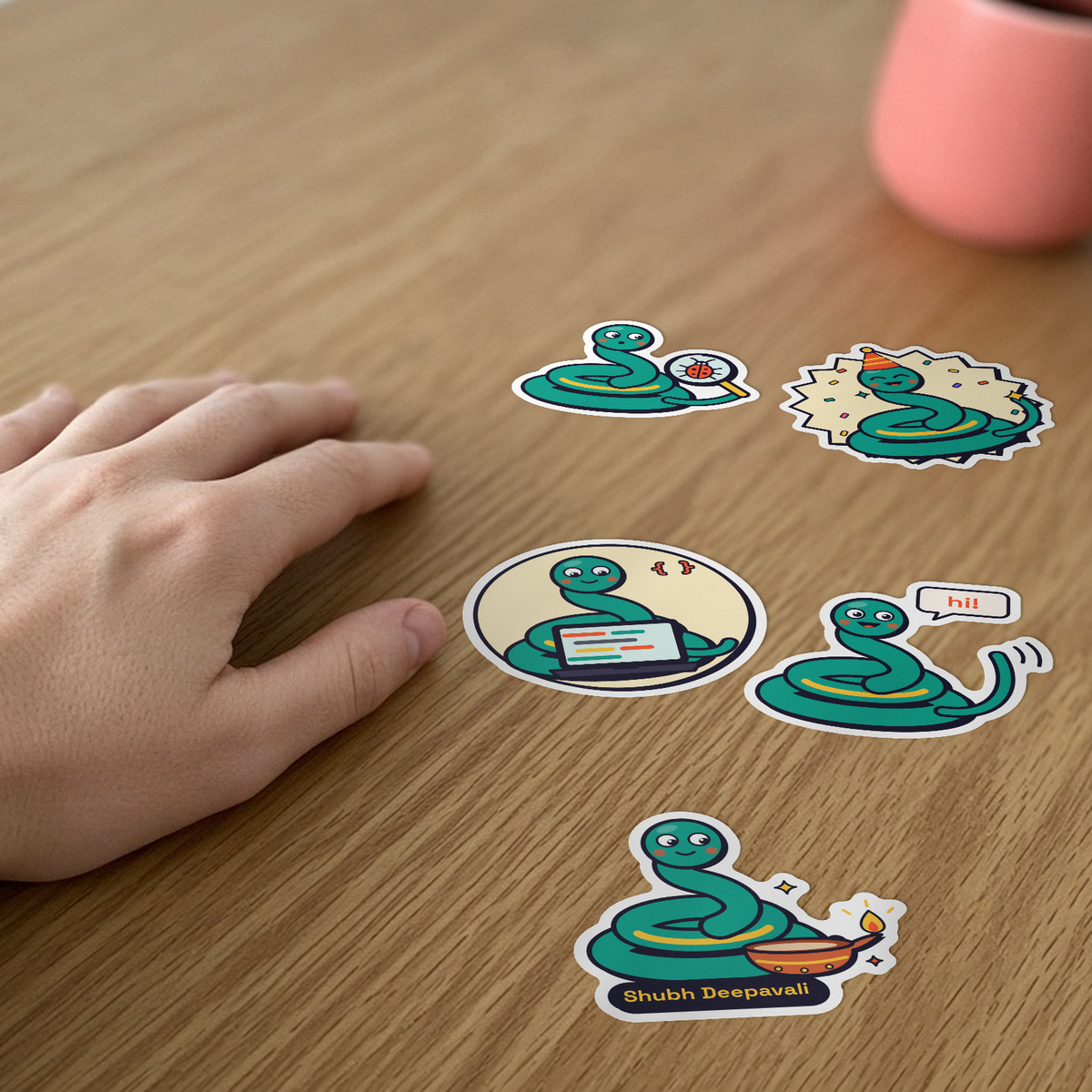 Pip Sticker Pack: five Pip the snake stickers, including Pip coding and Pip lighting a diya, on a desk beside a hand.
