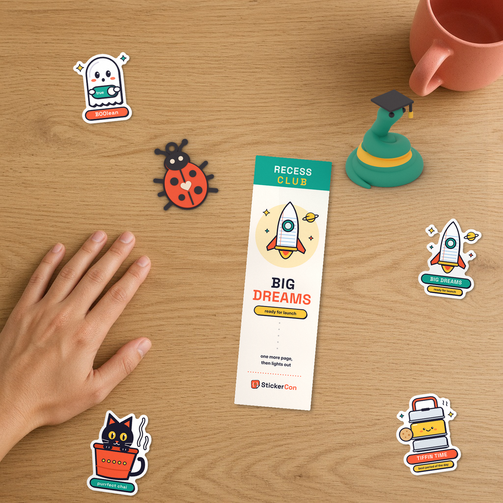 Top-down studio flat-lay on an oak desk showing the Recess Club Kids Set beside a hand for scale: Big Dreams rocket bookmark front, 3D-printed Graduate Pip, 3D-printed Bug Keychain, and Tiffin Time, Big Dreams, Kulhad Cat, and BOOlean Ghost stickers