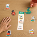 Top-down studio flat-lay on an oak desk showing the Recess Club Kids Set beside a hand for scale: Big Dreams rocket bookmark front, 3D-printed Graduate Pip, 3D-printed Bug Keychain, and Tiffin Time, Big Dreams, Kulhad Cat, and BOOlean Ghost stickers