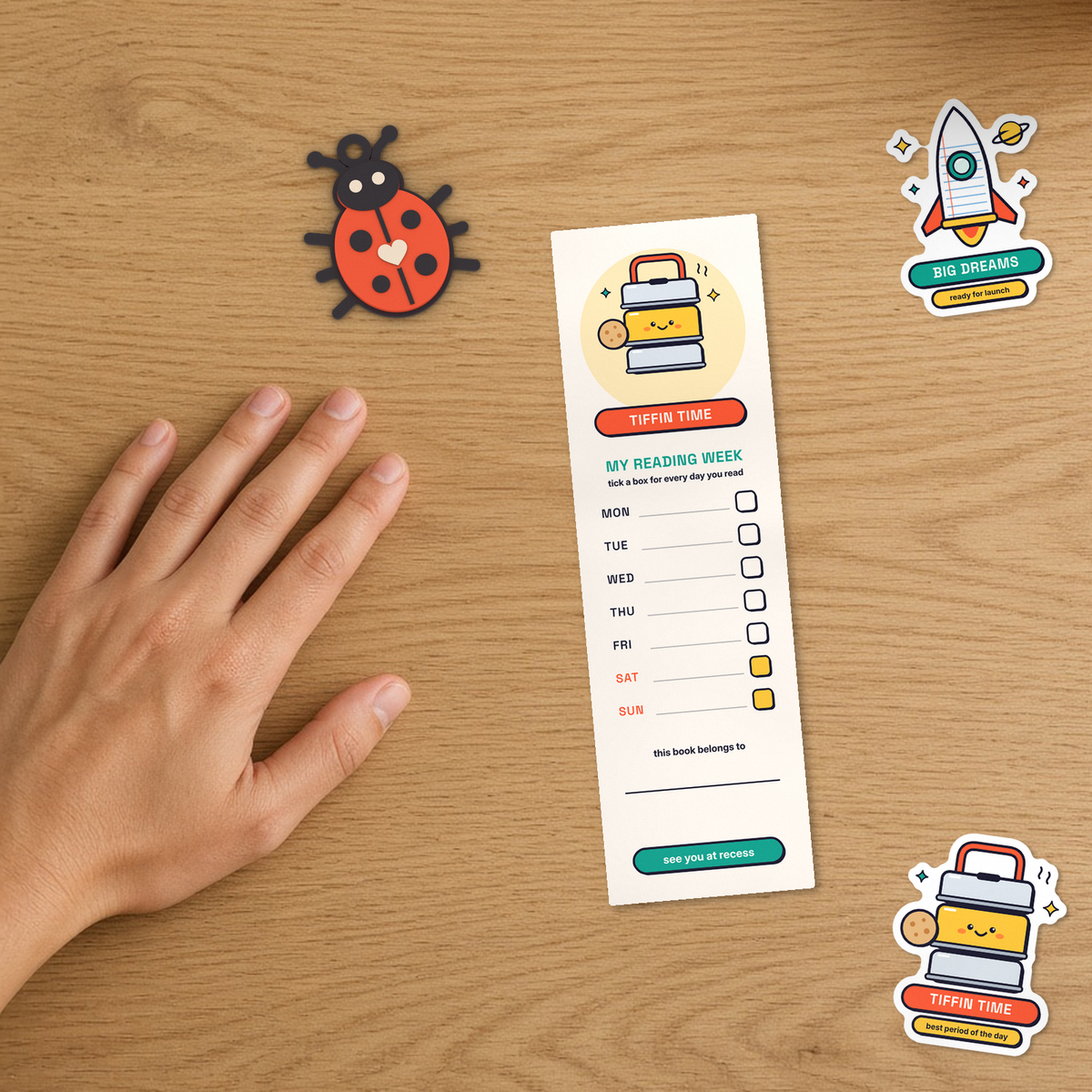 Oak desk flat-lay showing the back of the Recess Club bookmark with the My Reading Week Monday-to-Sunday tick tracker alongside the 3D-printed Bug Keychain and Big Dreams and Tiffin Time vinyl stickers beside a hand for scale