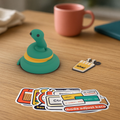 StickerCon Starter Bundle: classic Pip the snake, a chai keychain and the ten Desi Dev stickers fanned out on a wooden desk.