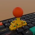 Macro photo of the 3D-printed articulated 4-hinge WASD Arcade Stick Keycap Rig with yellow keycap base blocks, teal hinge pins and a coral ball-top on a dark mechanical keyboard on an oak desk